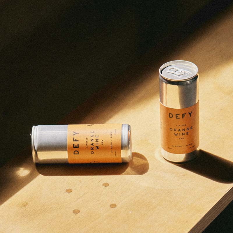 A DEFY orange wine can upright next to a can on its side on a wooden shelf