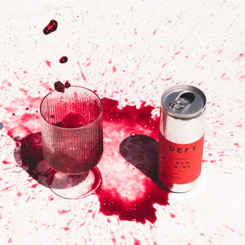 Messy red wine dropping into glass next to DEFY can of red wine.