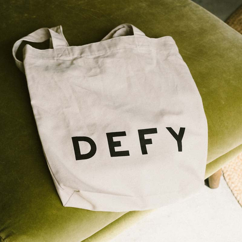 DEFY recycled cotton tote bag with the DEFY wordmark, on a green sofa.